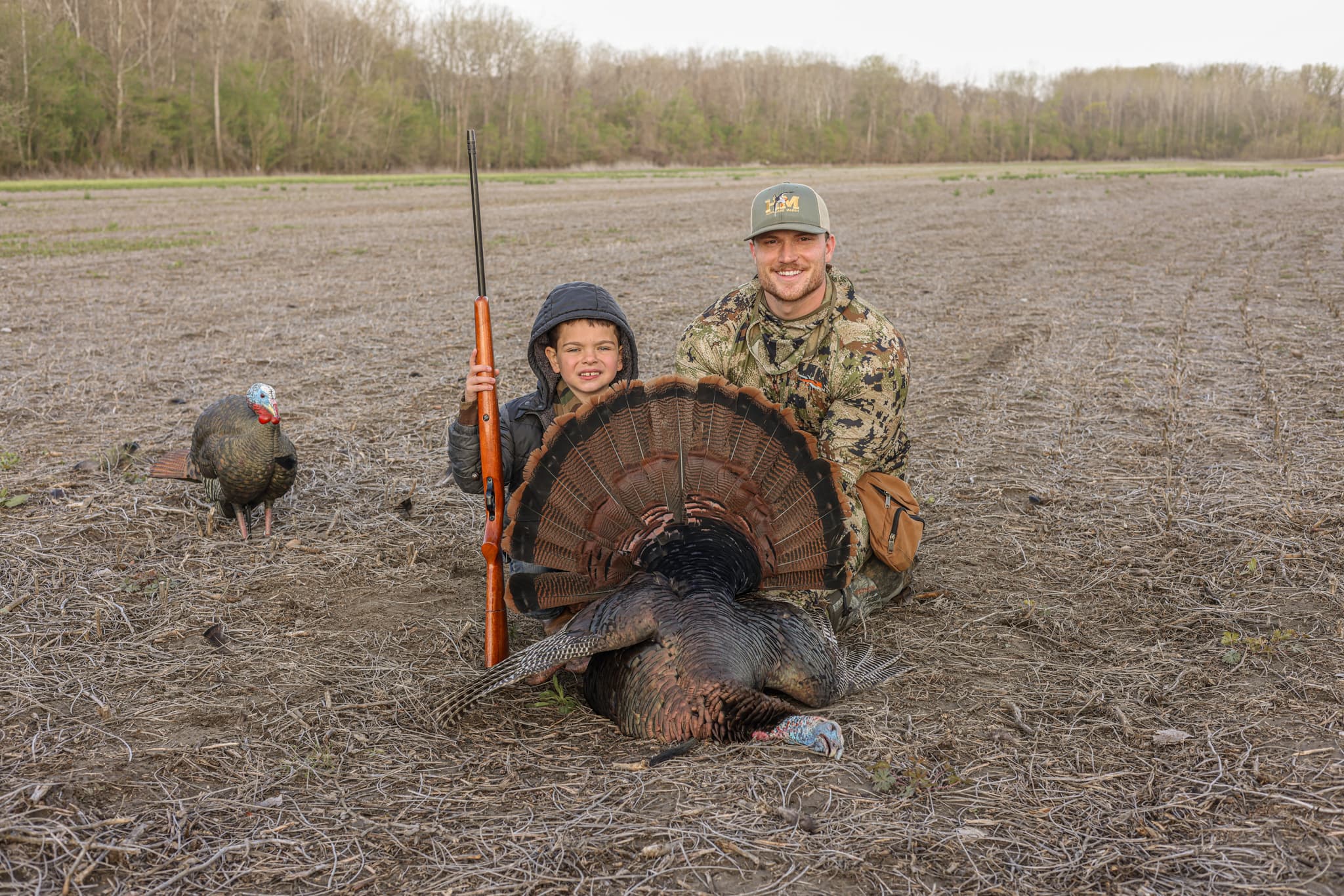 Two hunters, one young, kneeling with a spring gobbler fanned out in a field below the timber